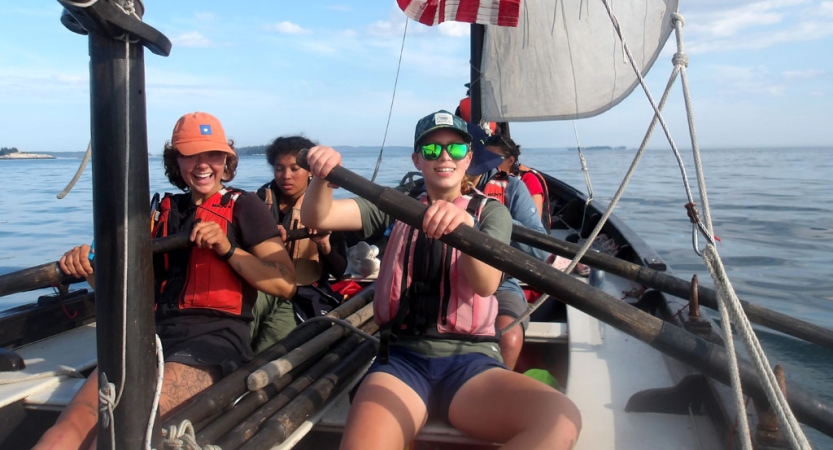 People wearing life jackets use the oars of a sailboat and smile for the photo. 